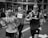 A group of runners participating in a city marathon or running event. They are captured mid-stride with expressions of focus and determination. The background features spectators and urban structures, indicative of a street race.