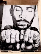A black and white mural of a man's face with closed eyes, displaying an expression of serenity. His hands are in front, showing tattoos on the knuckles that read 'JOY' and 'PAIN'. The words 'FROM DETROIT' are vertically written beside the hands.