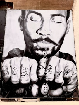 A black and white mural of a man's face with closed eyes, displaying an expression of serenity. His hands are in front, showing tattoos on the knuckles that read 'JOY' and 'PAIN'. The words 'FROM DETROIT' are vertically written beside the hands.