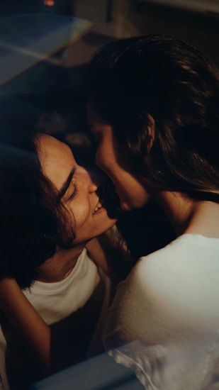 Two people are intimately close, with one gently smiling at the other. There's a soft, natural light illuminating their faces, creating a warm and tender atmosphere.