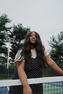 A person with long dark hair and a polka dot dress stands at a tennis net, with trees and a cloudy sky in the background. A tattoo of a butterfly is visible on their arm. The setting appears to be an outdoor tennis court.