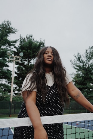 A person with long dark hair and a polka dot dress stands at a tennis net, with trees and a cloudy sky in the background. A tattoo of a butterfly is visible on their arm. The setting appears to be an outdoor tennis court.