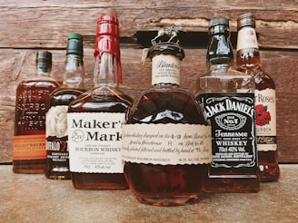 A collection of various whiskey bottles arranged on a wooden surface. The labels display brands like Maker's Mark, Jack Daniel's, and Blanton's. Each bottle has distinct label designs and unique bottle shapes, with some having wax seals or intricate tops.
