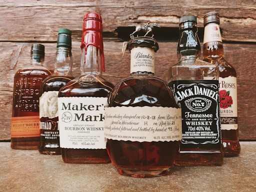 A collection of various whiskey bottles arranged on a wooden surface. The labels display brands like Maker's Mark, Jack Daniel's, and Blanton's. Each bottle has distinct label designs and unique bottle shapes, with some having wax seals or intricate tops.