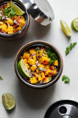A colorful salsa made with chopped mangoes, red onions, red bell peppers, and cilantro, garnished with lime wedges and placed in a stainless steel container with its lid partially open. Another container sits next to it and additional lime wedges and cilantro leaves are scattered around.