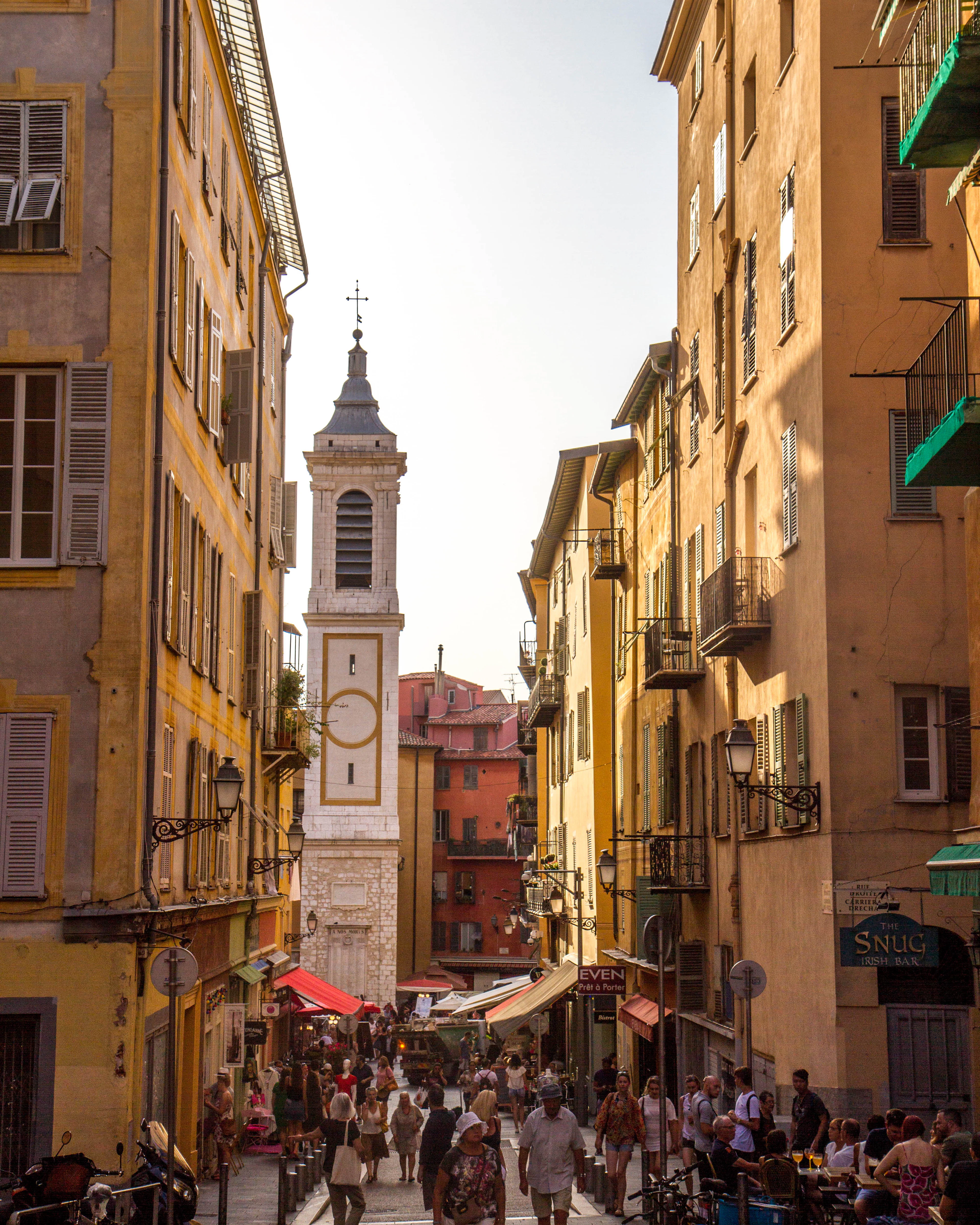 Late golden light down a busy street in Vieux Nice, ochre and yellow shuttered facades on both sides, market awnings and a bar terrace at the far end, and the white clock tower of the old town standing above the crowd.