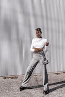A person stands in front of a corrugated metal wall wearing a white long-sleeve crop top, gray plaid high-waisted pants, and black shoes. A small black purse with a chain strap hangs at their side, and they are adorned with layered necklaces. The ground is paved with small stones, and there's a casual, confident pose.