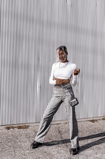 A person stands in front of a corrugated metal wall wearing a white long-sleeve crop top, gray plaid high-waisted pants, and black shoes. A small black purse with a chain strap hangs at their side, and they are adorned with layered necklaces. The ground is paved with small stones, and there's a casual, confident pose.