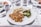 A white plate displays a meal consisting of roasted Brussels sprouts, cauliflower, and a serving of pasta casserole topped with breadcrumbs. The table around the plate holds a cup of black coffee, a glass of water, and additional dishes slightly out of focus.