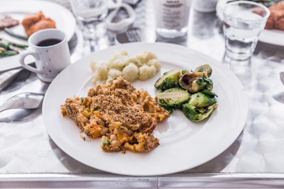 A white plate displays a meal consisting of roasted Brussels sprouts, cauliflower, and a serving of pasta casserole topped with breadcrumbs. The table around the plate holds a cup of black coffee, a glass of water, and additional dishes slightly out of focus.
