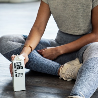 A person sitting cross-legged on a wooden floor while holding a carton labeled 'Boxed Water is Better.' They are wearing a grey short-sleeve top, grey leggings, and off-white sneakers.
