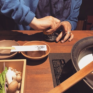 A person is squeezing a citrus fruit using a metal juicer. Two small bowls and a pair of chopsticks are placed on a wooden table. Fresh ingredients, including mushrooms and greens, are visible in a rectangular wooden container. A pot filled with a white liquid and a wooden spatula is also present on an electric stovetop.