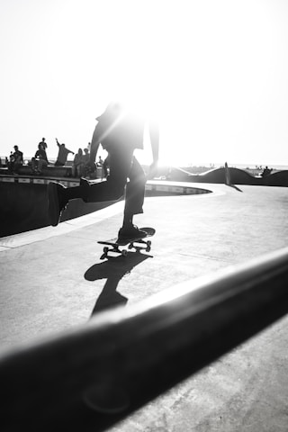 A silhouette of a person performing a trick on a skateboard, captured in motion against a bright sunlit background. The image is in black and white, highlighting the contrast between the skateboarder, their shadow, and the smooth surface of the skatepark. In the background, several people are visible, enhancing the dynamic and lively atmosphere.