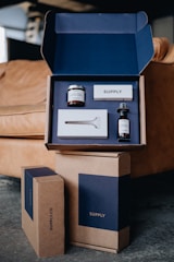 A sleek and neatly organized set of grooming products displayed in an open blue and brown packaging box. The set includes a jar of shaving cream, a razor, a bottle of post-shave tonic, and a soap bar. The packaging and products have a modern, minimalist design, with the brand name 'SUPPLY' prominently displayed. The setting is a cozy interior with a leather couch in the background.