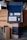 A sleek and neatly organized set of grooming products displayed in an open blue and brown packaging box. The set includes a jar of shaving cream, a razor, a bottle of post-shave tonic, and a soap bar. The packaging and products have a modern, minimalist design, with the brand name 'SUPPLY' prominently displayed. The setting is a cozy interior with a leather couch in the background.
