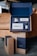 A sleek and neatly organized set of grooming products displayed in an open blue and brown packaging box. The set includes a jar of shaving cream, a razor, a bottle of post-shave tonic, and a soap bar. The packaging and products have a modern, minimalist design, with the brand name 'SUPPLY' prominently displayed. The setting is a cozy interior with a leather couch in the background.