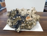 A large mineral specimen with clusters of dark, blackish rocks interspersed with shiny pyrite crystals and white quartz. The specimen sits on a wooden surface with a white paper underneath. Various textures are visible, including rough, jagged edges and smoother, rounded mineral formations.