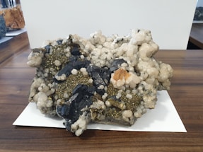 A large mineral specimen with clusters of dark, blackish rocks interspersed with shiny pyrite crystals and white quartz. The specimen sits on a wooden surface with a white paper underneath. Various textures are visible, including rough, jagged edges and smoother, rounded mineral formations.