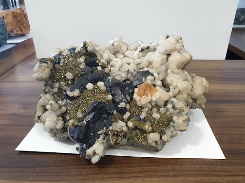 A large mineral specimen with clusters of dark, blackish rocks interspersed with shiny pyrite crystals and white quartz. The specimen sits on a wooden surface with a white paper underneath. Various textures are visible, including rough, jagged edges and smoother, rounded mineral formations.