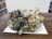 A large mineral specimen with clusters of dark, blackish rocks interspersed with shiny pyrite crystals and white quartz. The specimen sits on a wooden surface with a white paper underneath. Various textures are visible, including rough, jagged edges and smoother, rounded mineral formations.