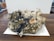 A large mineral specimen with clusters of dark, blackish rocks interspersed with shiny pyrite crystals and white quartz. The specimen sits on a wooden surface with a white paper underneath. Various textures are visible, including rough, jagged edges and smoother, rounded mineral formations.