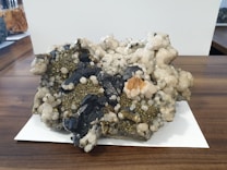 A large mineral specimen with clusters of dark, blackish rocks interspersed with shiny pyrite crystals and white quartz. The specimen sits on a wooden surface with a white paper underneath. Various textures are visible, including rough, jagged edges and smoother, rounded mineral formations.