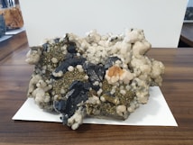 A large mineral specimen with clusters of dark, blackish rocks interspersed with shiny pyrite crystals and white quartz. The specimen sits on a wooden surface with a white paper underneath. Various textures are visible, including rough, jagged edges and smoother, rounded mineral formations.