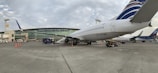 A commercial airplane is parked on the tarmac near an airport terminal. The terminal building has large glass windows and several signs, including one that reads 'Aeropuerto de Guayaquil José Joaquín de Olmedo.' Various ground service vehicles and equipment are visible around the aircraft, such as baggage carts and cones marking off areas. Partly cloudy skies are present above.