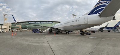 A commercial airplane is parked on the tarmac near an airport terminal. The terminal building has large glass windows and several signs, including one that reads 'Aeropuerto de Guayaquil José Joaquín de Olmedo.' Various ground service vehicles and equipment are visible around the aircraft, such as baggage carts and cones marking off areas. Partly cloudy skies are present above.