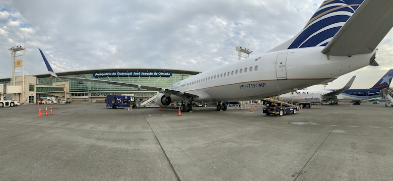 A commercial airplane is parked on the tarmac near an airport terminal. The terminal building has large glass windows and several signs, including one that reads 'Aeropuerto de Guayaquil José Joaquín de Olmedo.' Various ground service vehicles and equipment are visible around the aircraft, such as baggage carts and cones marking off areas. Partly cloudy skies are present above.