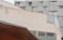 An urban architectural scene featuring a modern building with geometric shapes and patterns. The structure includes large glass windows and metal siding, with balconies on one side. The image captures a section of the building with a blend of concrete and glass materials.