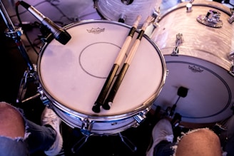 A close-up view of a drum set, featuring a snare drum with light-colored drumsticks resting on it. A microphone is positioned near the drum, indicating a setup for recording or performance. The bass drum and other parts of the drum kit are visible in the background. The scene includes a musician's legs clad in torn jeans and sneakers, suggesting a casual or informal setting.