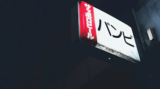 A rectangular illuminated sign featuring Japanese characters is mounted against a dark background. The sign has a red vertical panel on the left side with white text and a larger white section with black text on the main area.