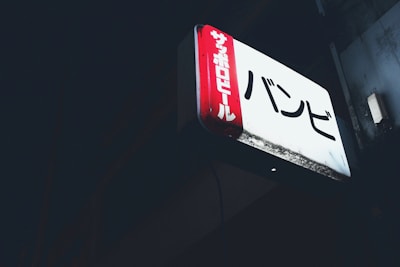 A rectangular illuminated sign featuring Japanese characters is mounted against a dark background. The sign has a red vertical panel on the left side with white text and a larger white section with black text on the main area.