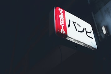 A rectangular illuminated sign featuring Japanese characters is mounted against a dark background. The sign has a red vertical panel on the left side with white text and a larger white section with black text on the main area.