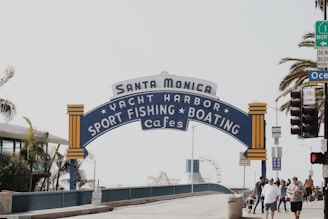 A large, iconic sign arching over a walkway displays the words 'Santa Monica Yacht Harbor Sport Fishing Boating Cafes'. The sign features blue and yellow accents and stands in front of palm trees and a clear sky. A group of people walks beneath the sign, with some traffic lights and a building visible on the side.