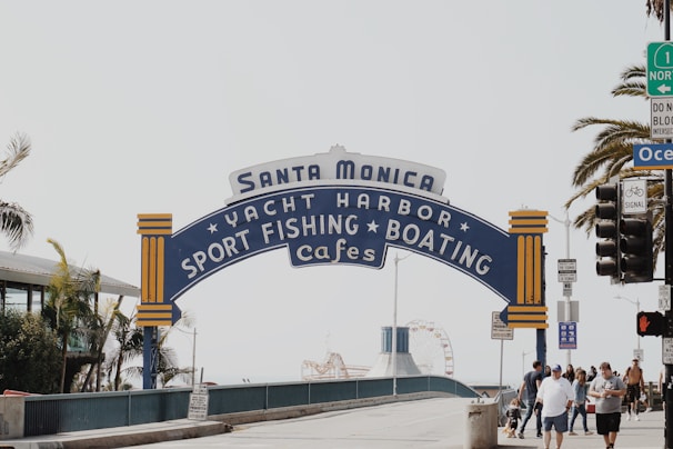 A large, iconic sign arching over a walkway displays the words 'Santa Monica Yacht Harbor Sport Fishing Boating Cafes'. The sign features blue and yellow accents and stands in front of palm trees and a clear sky. A group of people walks beneath the sign, with some traffic lights and a building visible on the side.