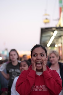 A woman in a red hoodie is expressing surprise or excitement, with her mouth open and hands on her cheeks. There are several people in the background walking in what appears to be a busy outdoor setting, likely a fair or festival, with blurred lights and structures visible.