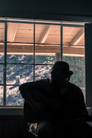 Musician playing guitar in front of a large window with natural light flooding the room.