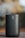 A cylindrical, portable speaker with a metallic mesh surface and a logo vertically aligned at the front. The background is blurred, highlighting the focus on the speaker, which has a sleek and modern design.