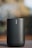 A cylindrical, portable speaker with a metallic mesh surface and a logo vertically aligned at the front. The background is blurred, highlighting the focus on the speaker, which has a sleek and modern design.