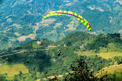 A colorful paraglider is soaring over lush, terraced green hills. The landscape is dotted with small houses and dense forest areas, with winding roads cutting through the scenery. The vibrant parachute stands out against the natural backdrop, creating a striking contrast.
