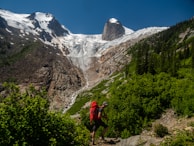 A person with a red backpack hikes through a lush green forest with a rocky mountain and snow-capped peaks in the background beneath a clear blue sky.