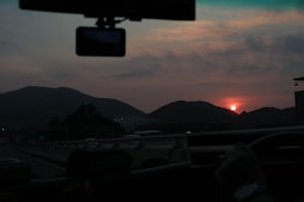 A view from inside a car driving along a highway during sunset. The landscape includes silhouetted mountains against a sky with scattered clouds and a setting sun glowing in an orange hue. The interior mirror and a navigation device or dashcam are visible inside the vehicle.