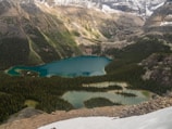 A serene landscape features a series of turquoise lakes nestled among dense green forests and surrounded by towering rocky mountains partially covered in snow. The natural beauty of the scene is enhanced by the lush greenery and diverse textures of the terrain.