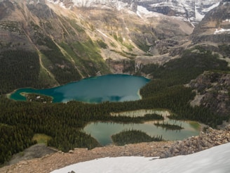 A serene landscape features a series of turquoise lakes nestled among dense green forests and surrounded by towering rocky mountains partially covered in snow. The natural beauty of the scene is enhanced by the lush greenery and diverse textures of the terrain.