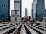 A modern urban scene featuring tall skyscrapers in the background and a cluster of parked trains in the foreground. The unique architecture of the buildings includes a honeycomb-like structure at the center. The image captures the busy atmosphere of a cityscape, with an emphasis on infrastructure and urban development.