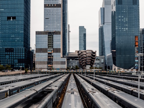 A modern urban scene featuring tall skyscrapers in the background and a cluster of parked trains in the foreground. The unique architecture of the buildings includes a honeycomb-like structure at the center. The image captures the busy atmosphere of a cityscape, with an emphasis on infrastructure and urban development.