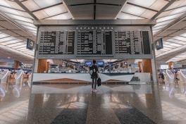 A modern airport terminal with a large departures board displaying various flight destinations and times. A person stands beneath the board, holding a bag, surrounded by sleek, futuristic check-in kiosks and reflective flooring.