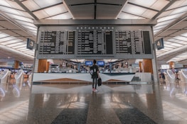 A modern airport terminal with a large departures board displaying various flight destinations and times. A person stands beneath the board, holding a bag, surrounded by sleek, futuristic check-in kiosks and reflective flooring.